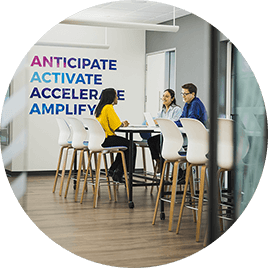 Three office employees work together on their laptops at a high-top table, in front of a sign that says "Anticipate; Activate; Accelerate; Amplify."