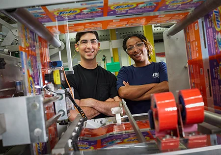 A male and female employee are in a manufacturing plant. They smile directly at the camera through some of the machinery.