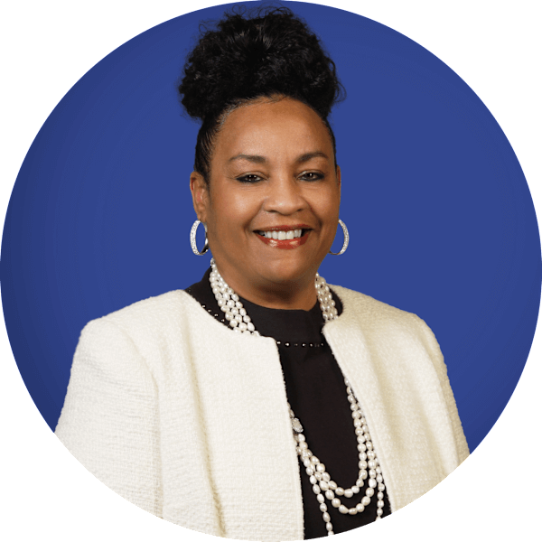 A Black woman with dark hair smiles at the camera. She wears a black shirt, white jacket, and pearl necklace.