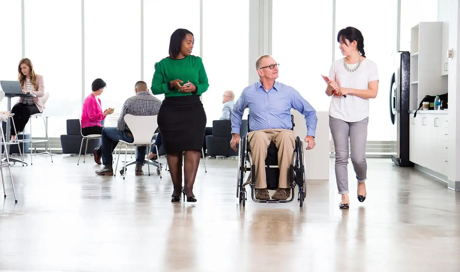 Two women walking beside a man in a wheelchair in an office environment