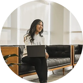 A woman with long, black hair sits on a bench with her laptop on her lap, listening intently to another colleague.