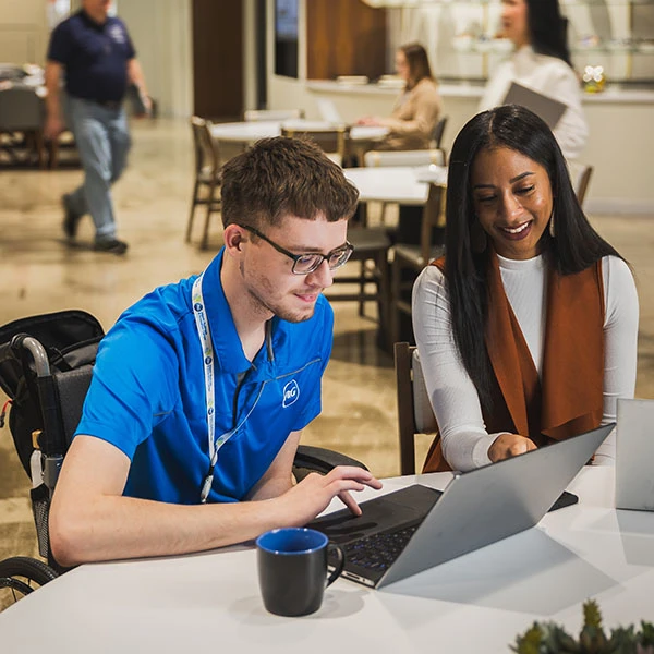 A man and woman sit together in an office and look at a laptop screen. Several other people are walking in the background.