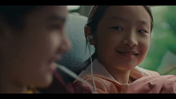 A young Asian girl on a school bus. She is smiling at another young girl who sits next to her, and they are sharing headphones.