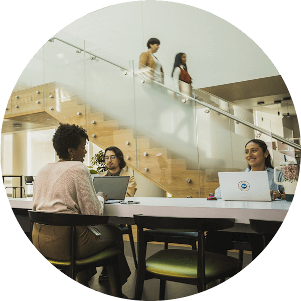 Several people sit together at various office work stations. Two women walk down a staircase in the background.