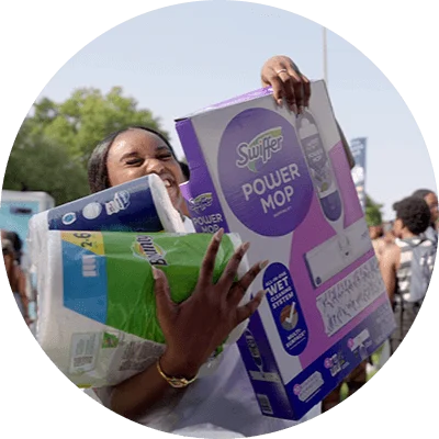 A young Black woman smiles as she carries an armful of household products, such as paper towels and toilet paper.