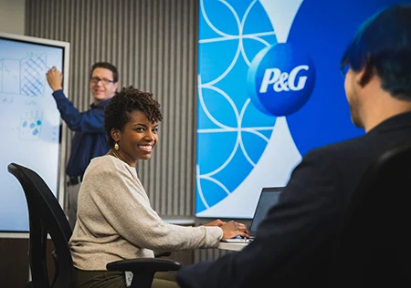 A male and female employee smile at and talk with one another while sitting at their desks at work, while a third employee writes on a white board in the background.