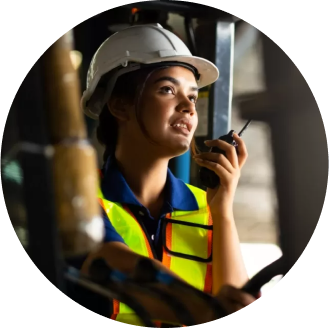 A  worker with a white hard hat and high-visibility safety vest talks into a walkie-talkie is in an industrial setting.