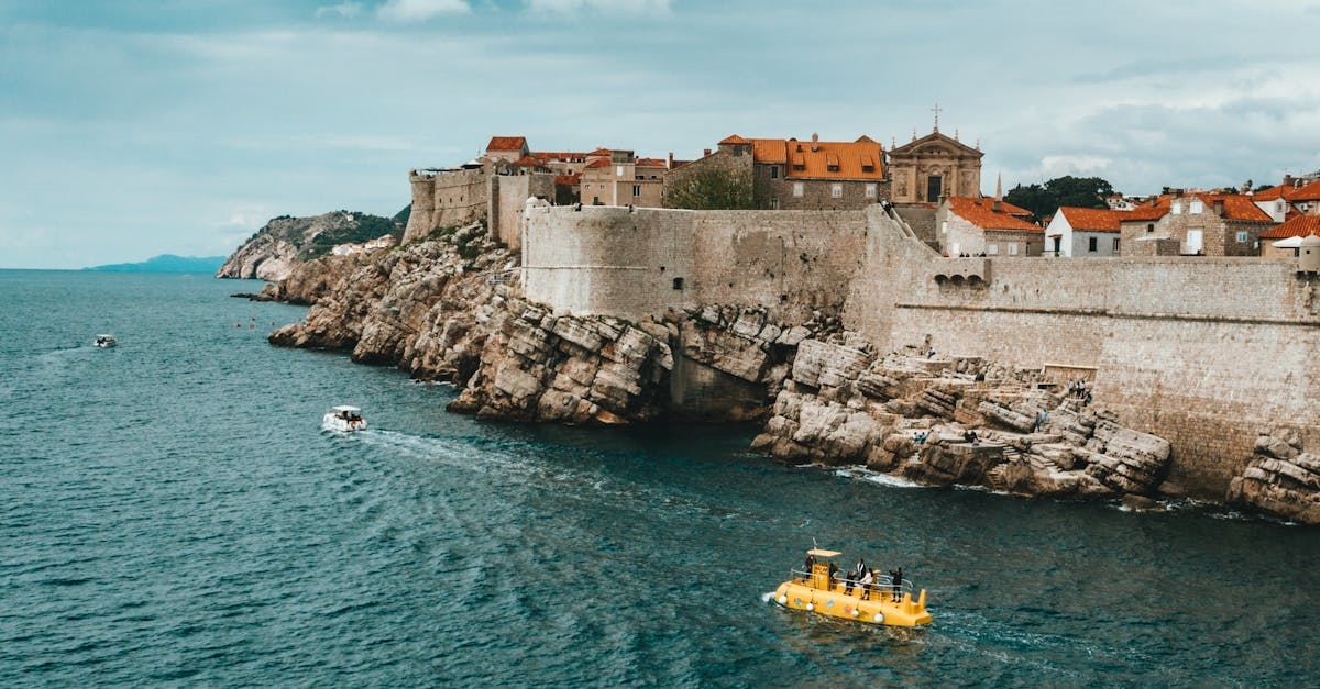 Modern boats floating on rippling sea near rocky coast of old town of Dubrovnik with historical buildings and ancient city walls