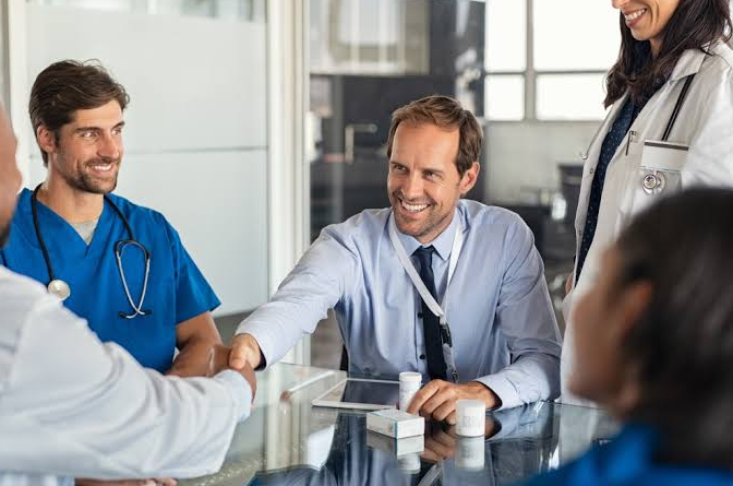 A medical device sales rep shaking hands with a surgeon while nurses and a practice administrator look on across a meeting table
