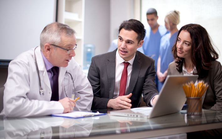 A physician, a sales rep and a practice manager reviewing information on a laptop together while clinical staff talk in the background