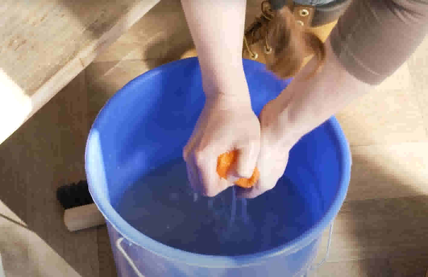 Handen knijpen een oranje spons uit boven een blauwe emmer water op een houten vloer.