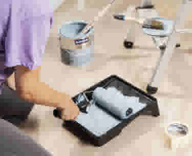 Persoon in paars shirt dompelt verfroller in verfbak, met verfblik en schildertape op de vloer tijdens renovatie.