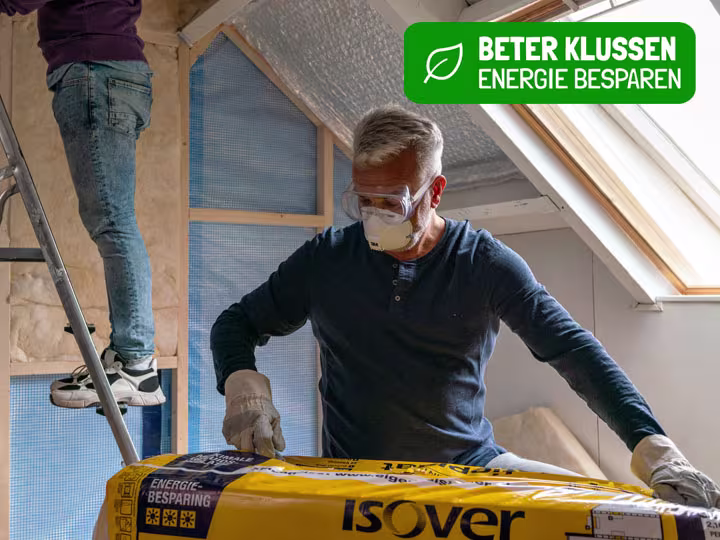 Man met veiligheidsbril en masker installeert isolatiemateriaal op zolder, met logo "Beter Klussen Energie Besparen"
