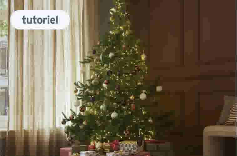Sapin de Noël décoré avec guirlandes lumineuses et boules colorées dans un salon avec rideaux beiges et cadeaux au pied.