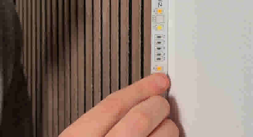 Een hand plaatst een LED-strip tegen een houten wand met verticale latten, naast een witte muur.