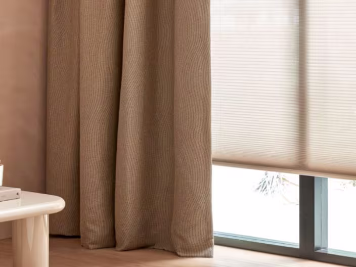 Bruine gordijnen naast crèmekleurige rolgordijnen voor een raam, met een glimp van een witte tafel aan de linkerkant.