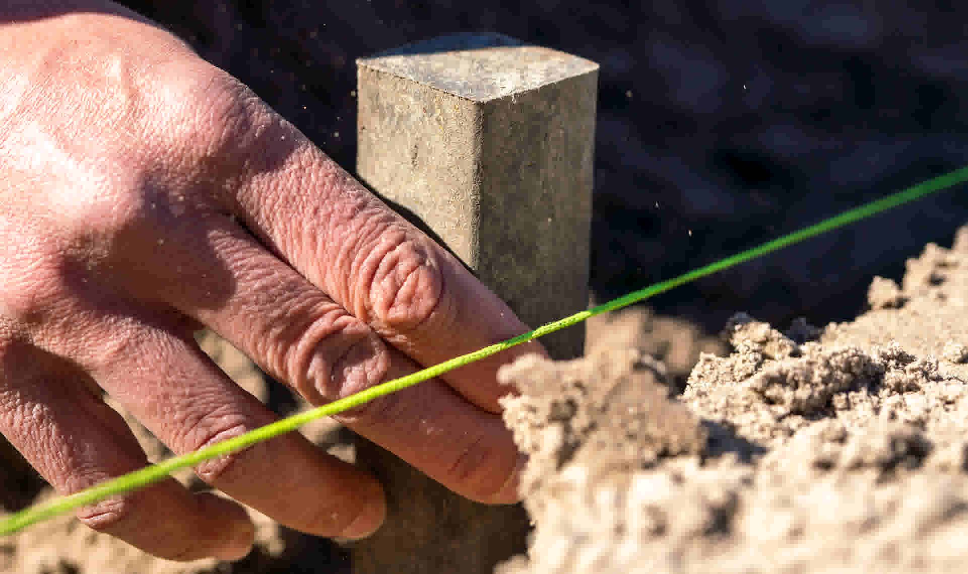 Hand houdt een groen koord strak tegen een houten paaltje, met zand zichtbaar op de achtergrond.