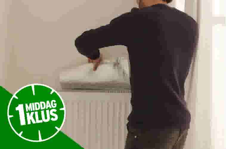 Man in zwarte trui schildert witte muur boven radiator met verfroller, met groene 1 Middag Klus badge.