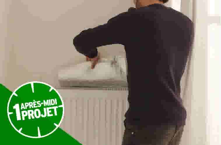 Homme en pull noir peignant un mur beige au-dessus d'un radiateur blanc avec logo vert "1 après-midi projet"
