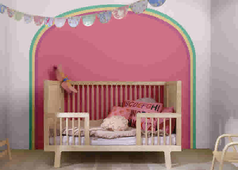 Lit bébé en bois avec coussins roses devant un mur rose orné d'une arche colorée et de guirlandes festives.