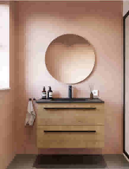 Moderne badkamer met houten wastafelmeubel, ronde spiegel en zalmroze muren. Zwarte wasbak en verzorgingsproducten zichtbaar.