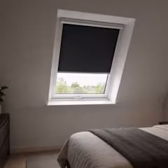 Store enrouleur noir occultant sur fenêtre de toit blanche dans une chambre lumineuse avec lit et commode sombre.