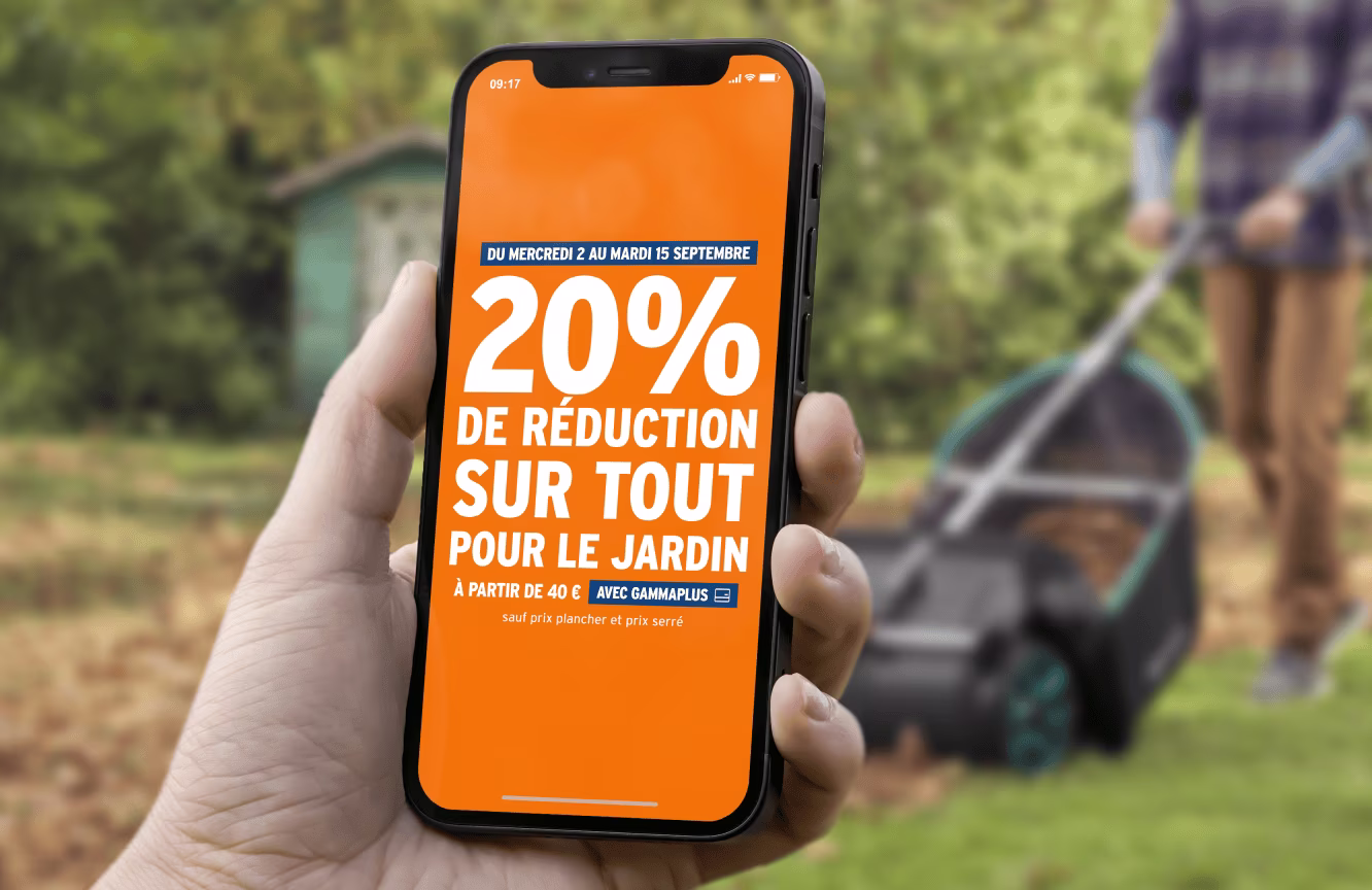 Main tenant un smartphone affichant une promotion orange de 20% de réduction sur tout pour le jardin dans un jardin.