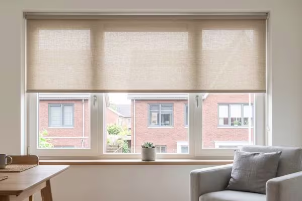Beige rolgordijnen halfopen voor een breed raam met uitzicht op bakstenen huizen. Lichte woonkamer met grijze stoel en houten tafel.