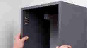 Hand plaatst een zwarte kolomkast tegen een witte muur, met de deur open.