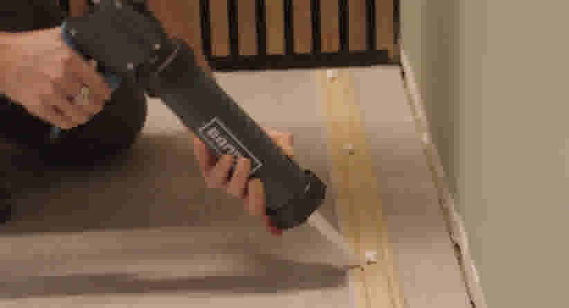 Persoon gebruikt een DeWalt kitpistool om kit aan te brengen langs een houten lat op een trap of vloer.