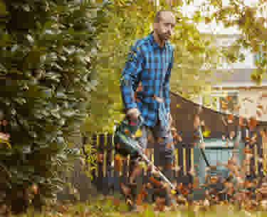 Jardinier en chemise à carreaux bleue arrosant des plantes dans un jardin automnal entouré de feuillage.