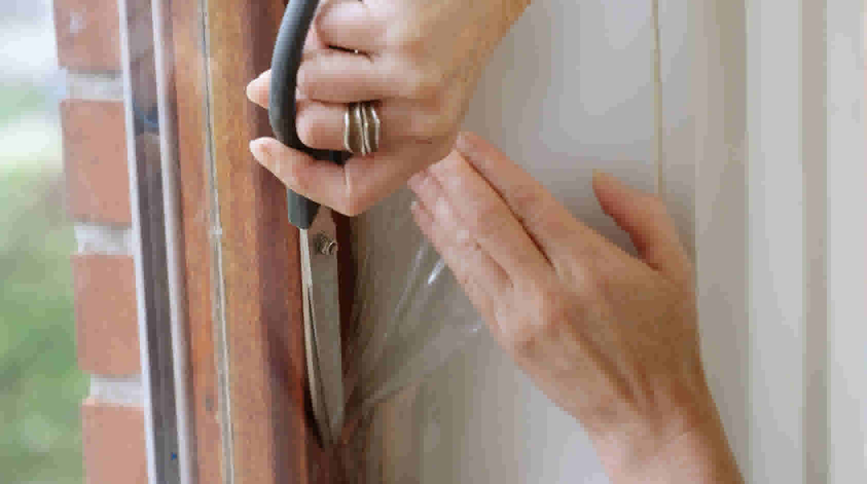 Handen plaatsen doorzichtig isolatiefolie op een houten raamkozijn met een speciaal gereedschap om tocht te voorkomen.