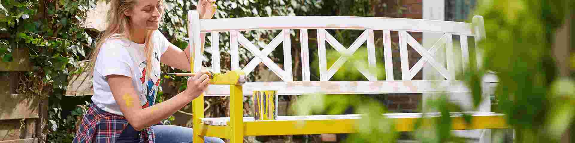 Une femme souriante peint un banc de jardin en jaune vif avec un pinceau, entourée de verdure.