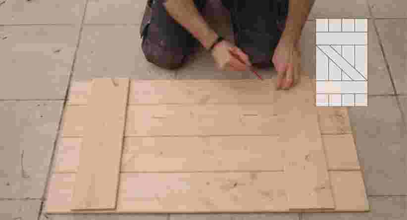 Persoon markeert houten planken op een tegelvloer tijdens het bouwen van een houten constructie, met een ontwerpschema zichtbaar.