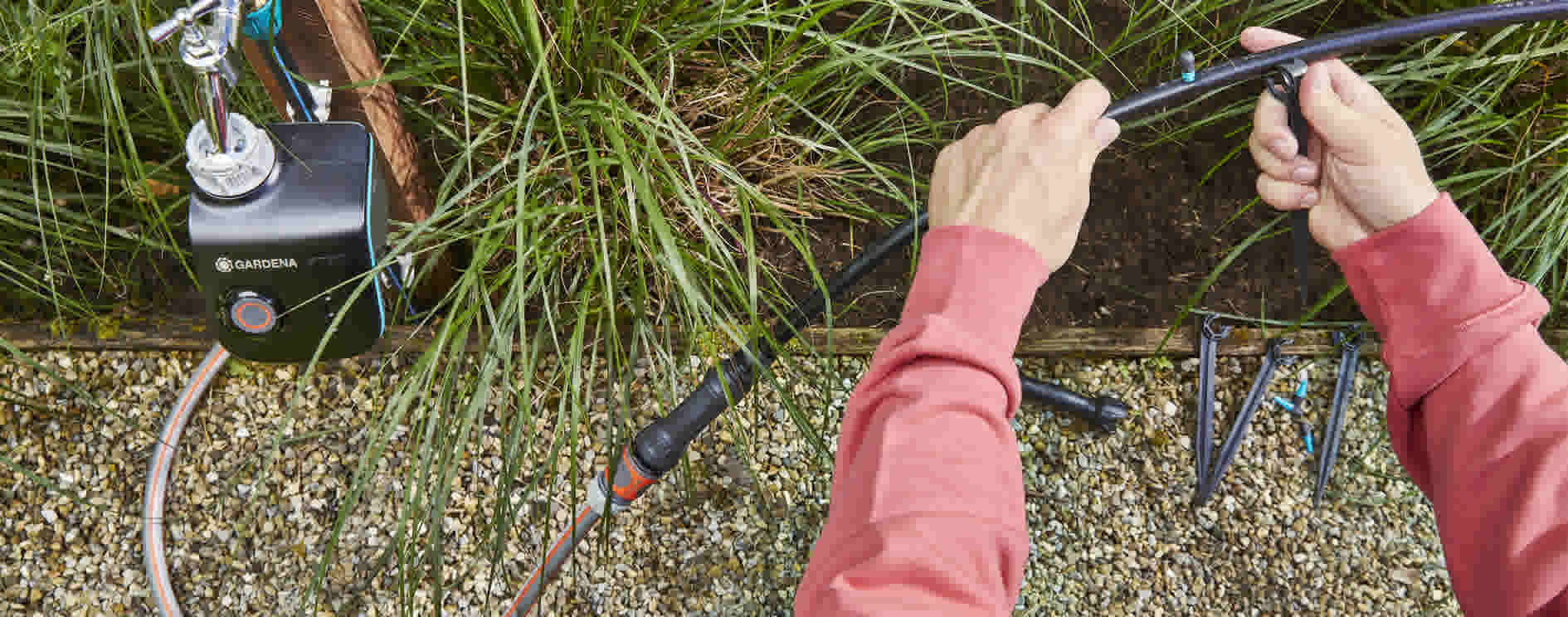 Mains en pull rose installant un système d'irrigation Gardena avec tuyaux noirs près d'une pelouse et zone de gravier.
