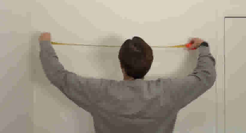 Persoon in grijze trui houdt een geel meetlint tegen een witte muur om een afstand op te meten.