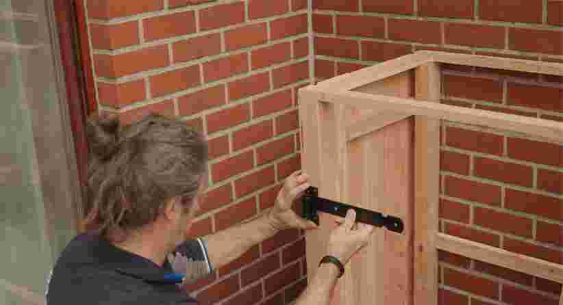 Persoon bevestigt een zwart metalen scharnier aan een houten frame tegen een bakstenen muur.