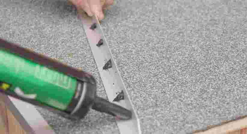 Hand plaatst een metalen strip met groene kitspuit op een grijs tapijt of vloerbedekking tijdens installatie.
