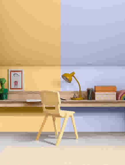 Kinderkamer met gele stoel voor houten bureau aan muur, geschilderd in geel en lila. Gele lamp, dozen en kunstwerk op bureau.
