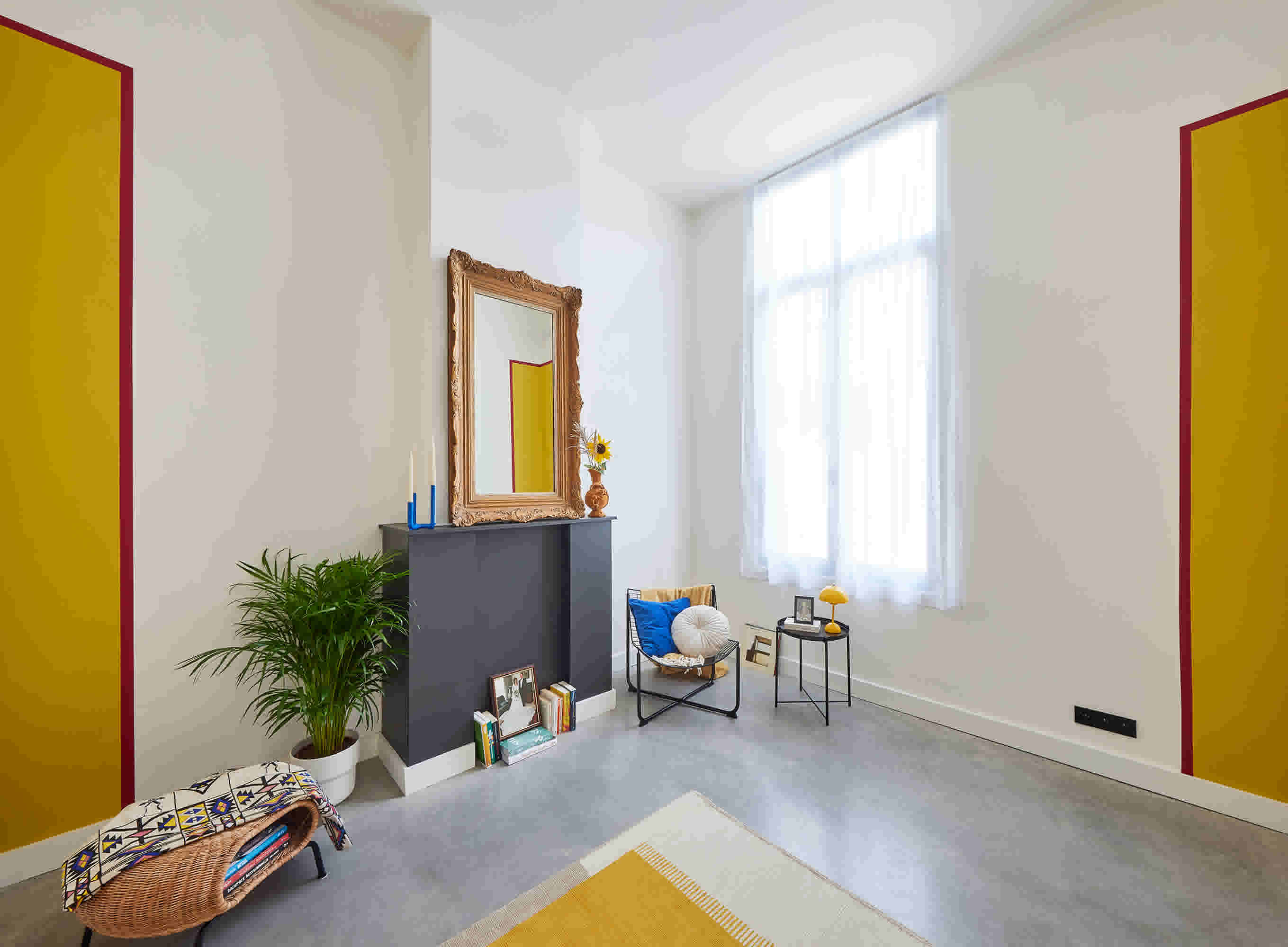 Moderne woonkamer met gele accenten, grijze schouw met spiegel, groene plant en stoel met blauwe kussen bij groot raam.