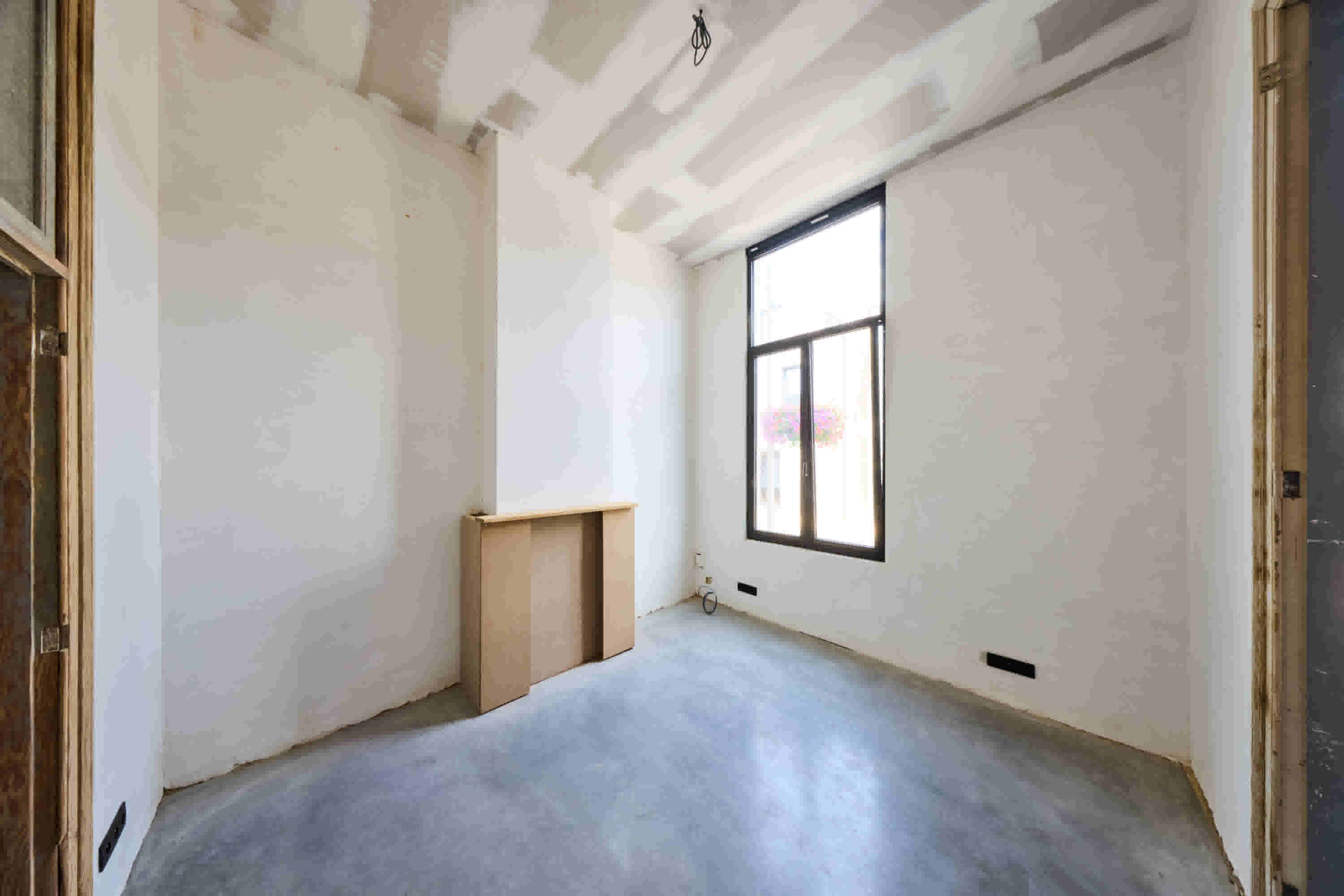 Lege kamer in renovatie met witte muren, betonnen vloer, groot raam en een houten kast tegen de muur.