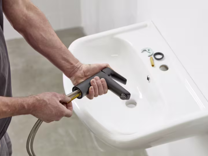 Une personne installe un nouveau robinet dans un lavabo blanc à l'aide d'outils. Quelques pièces sont posées sur le lavabo.