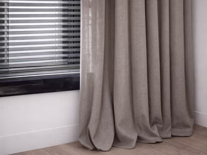 Rideau en tissu beige tombant près d'une fenêtre avec store vénitien gris, sur un parquet clair dans un intérieur moderne.