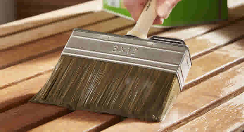 Hand houdt een brede kwast (8x12) vast en brengt beits aan op een houten oppervlak met planken.