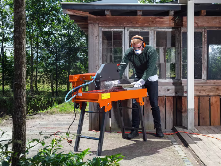 Man voor een schuur met mondkapje en gehoorbeschermers op staat buiten een zaagtafel te bedienen