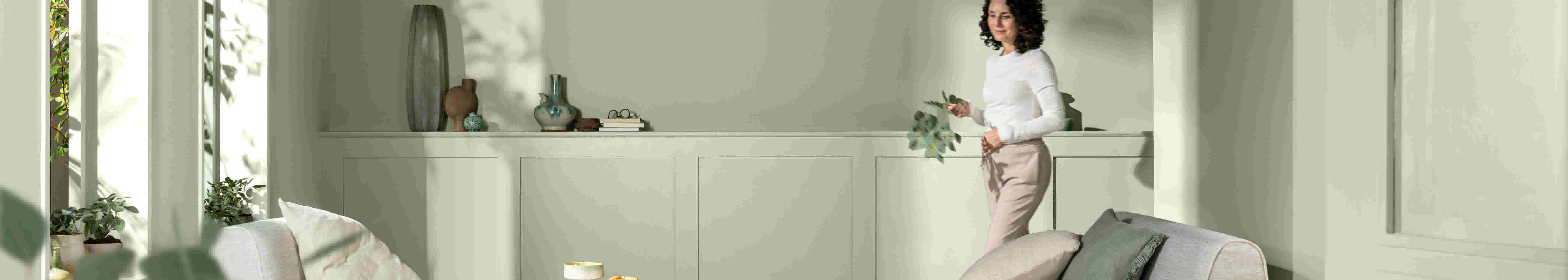 Vrouw in wit shirt en beige broek staat in lichte, moderne kamer met witte kasten en decoratieve vazen.