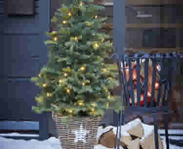 Petit sapin de Noël décoré de lumières dans un panier en osier, à côté d'un brasero et de bûches de bois.