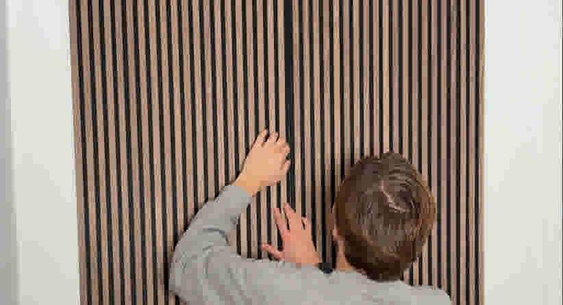 Persoon in grijze trui installeert een houten wandpaneel met verticale latten, gezien vanaf de achterkant.