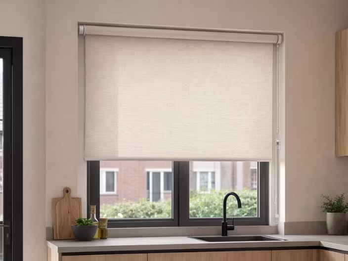 Beige rolgordijn voor een keukenraam boven een aanrecht met zwarte kraan, snijplank en kom met groenten.