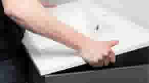 Hand plaatst een donkergrijze lade of opbergbak in een kastensysteem tegen een witte muur.